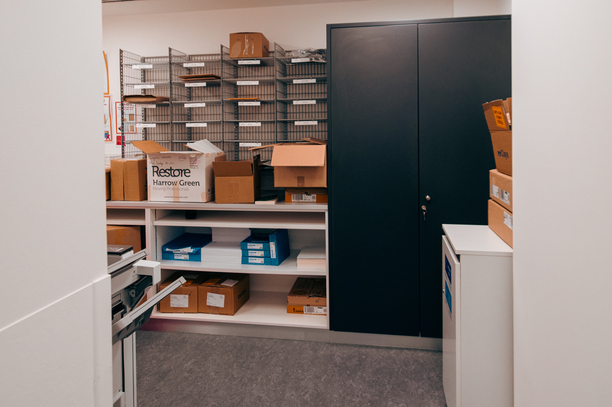 Printer room contains cubby holes for each team's post, cardboard boxes with stationery supplies, and large black storage cabinet.